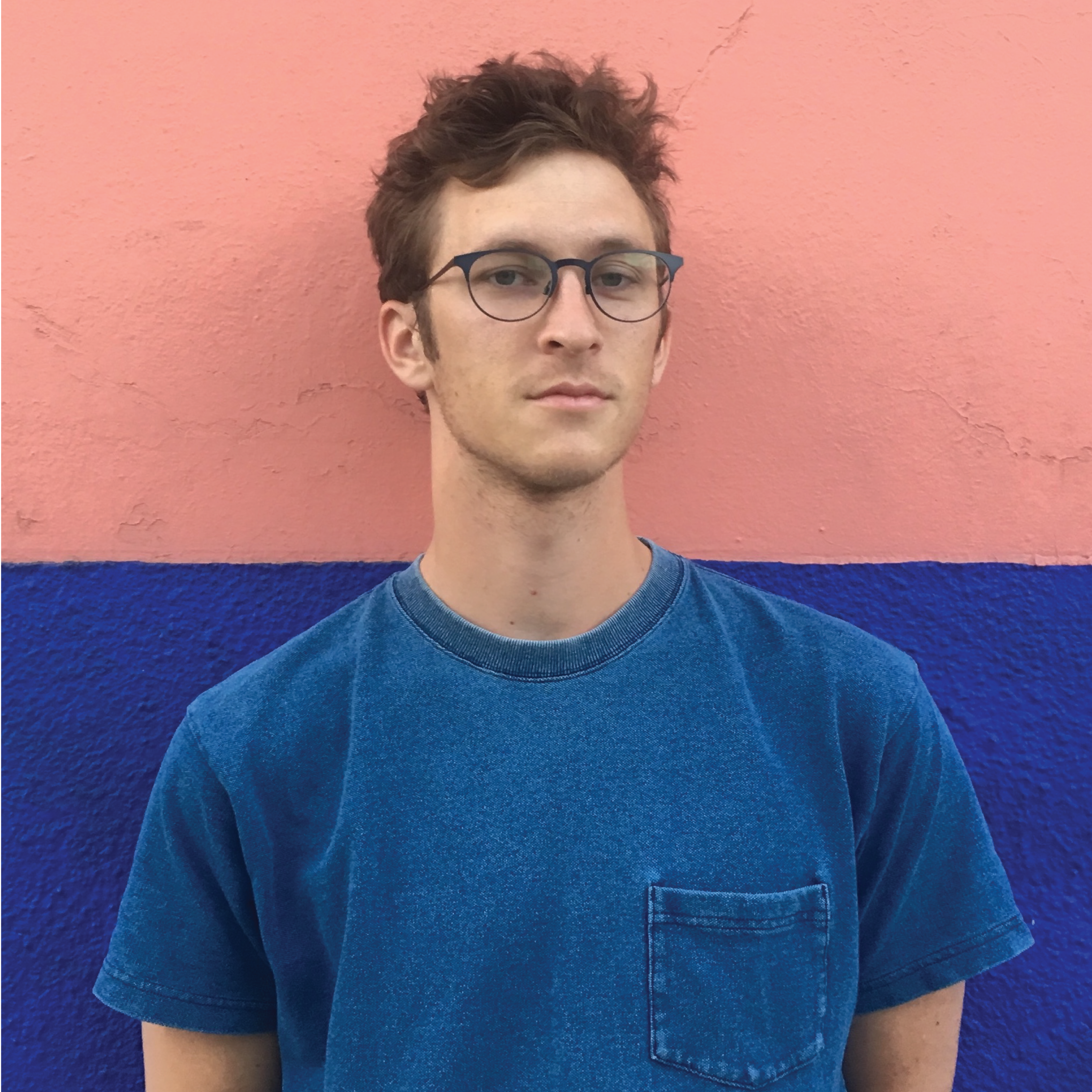 Portrait of Galen, a white man in his 20s with glasses and messy, short, brown hair. He is wearing a blue t-shirt and standing in front of a wall which is painted half blue and half pink.