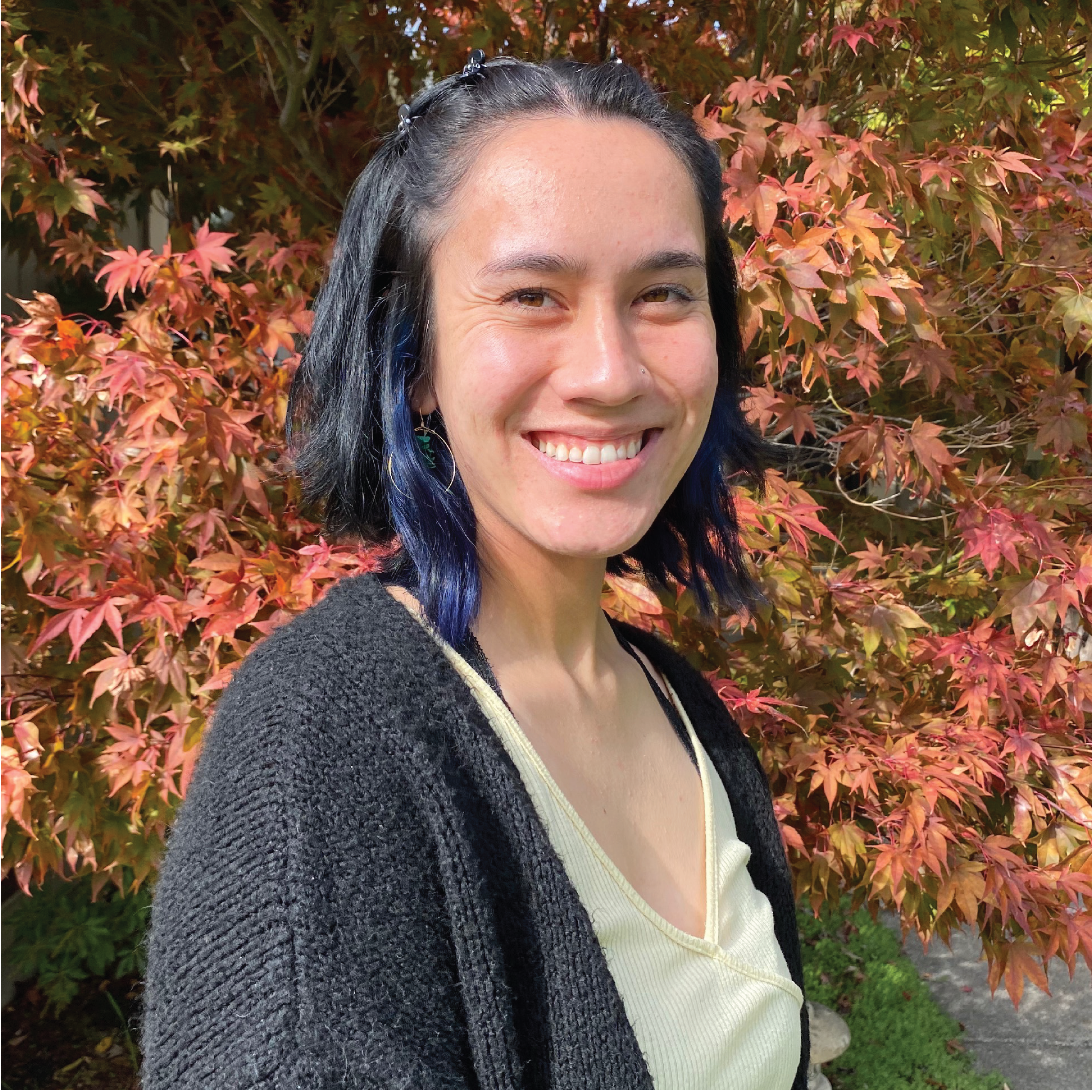 Young woman with shoulder-length dark hair wearing a yellow shirt and black cardigan standing in front of a maple tree smiling.
