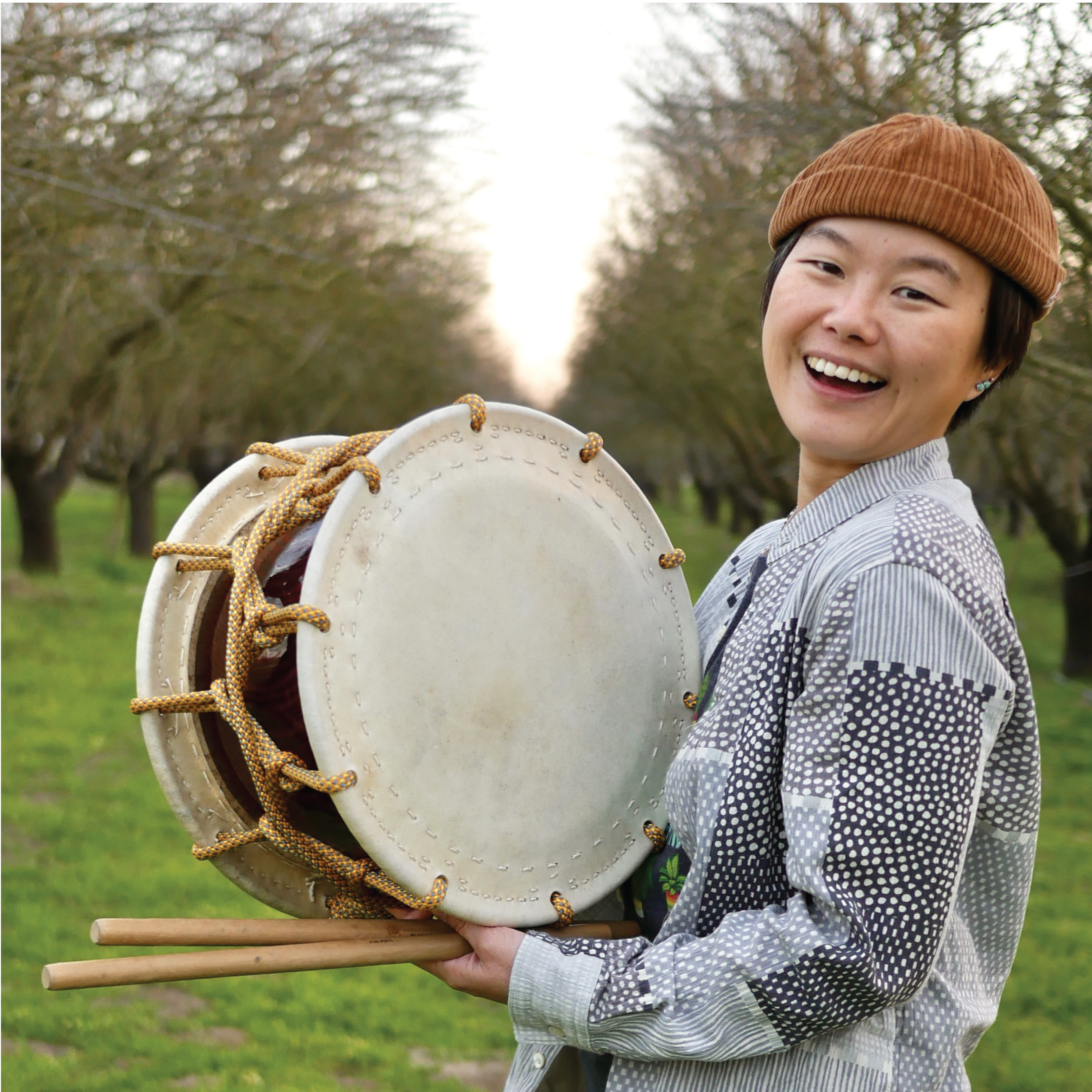 Portrait of ManMan, a short-haired Asian person with a brown beanie, wearing a light color patterned button down shirt holding a shime daiko on an almond tree farm.