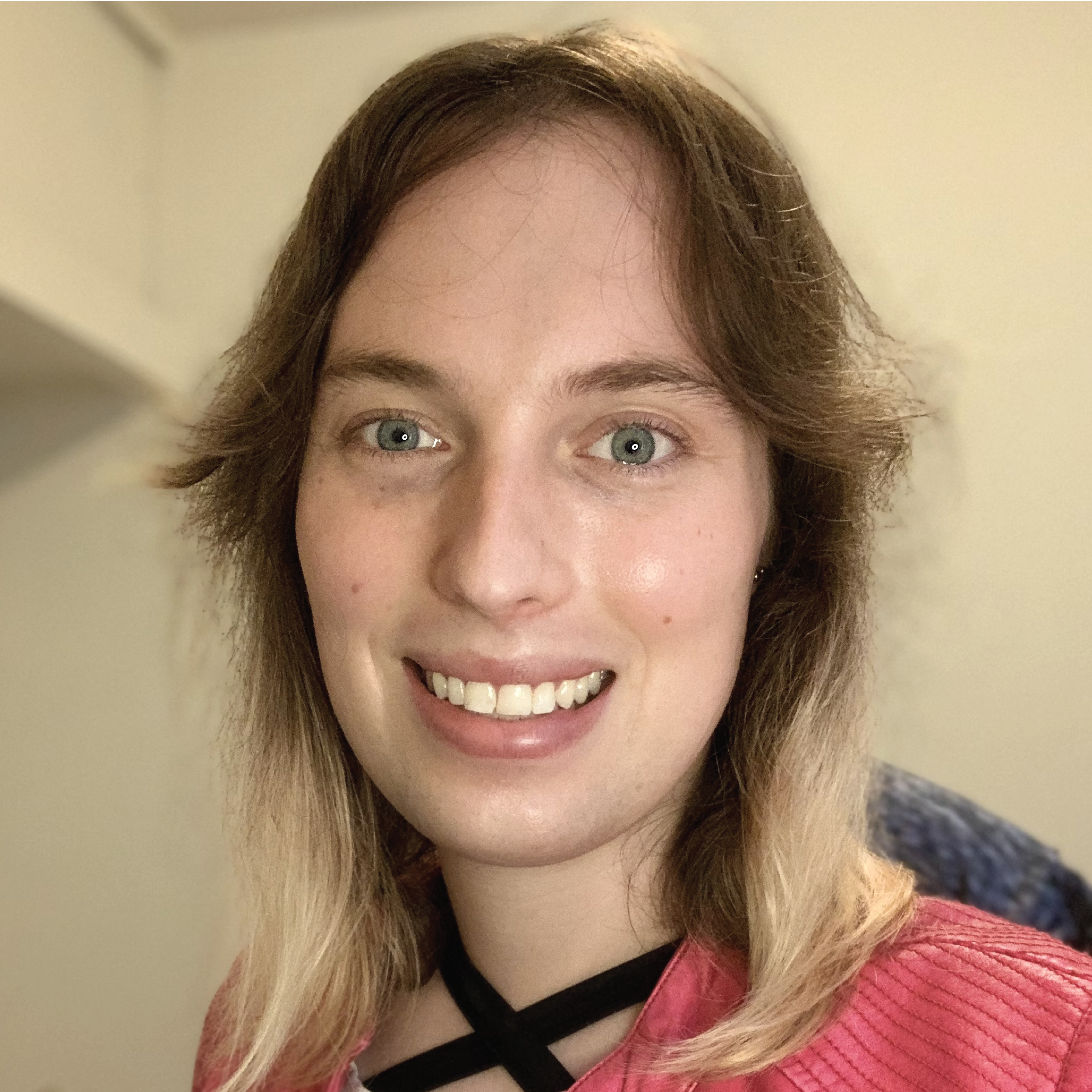 A white non-binary person with blue eyes and light brown hair with blond highlights wearing a pink jacket and a white shirt with black horizontal stripes