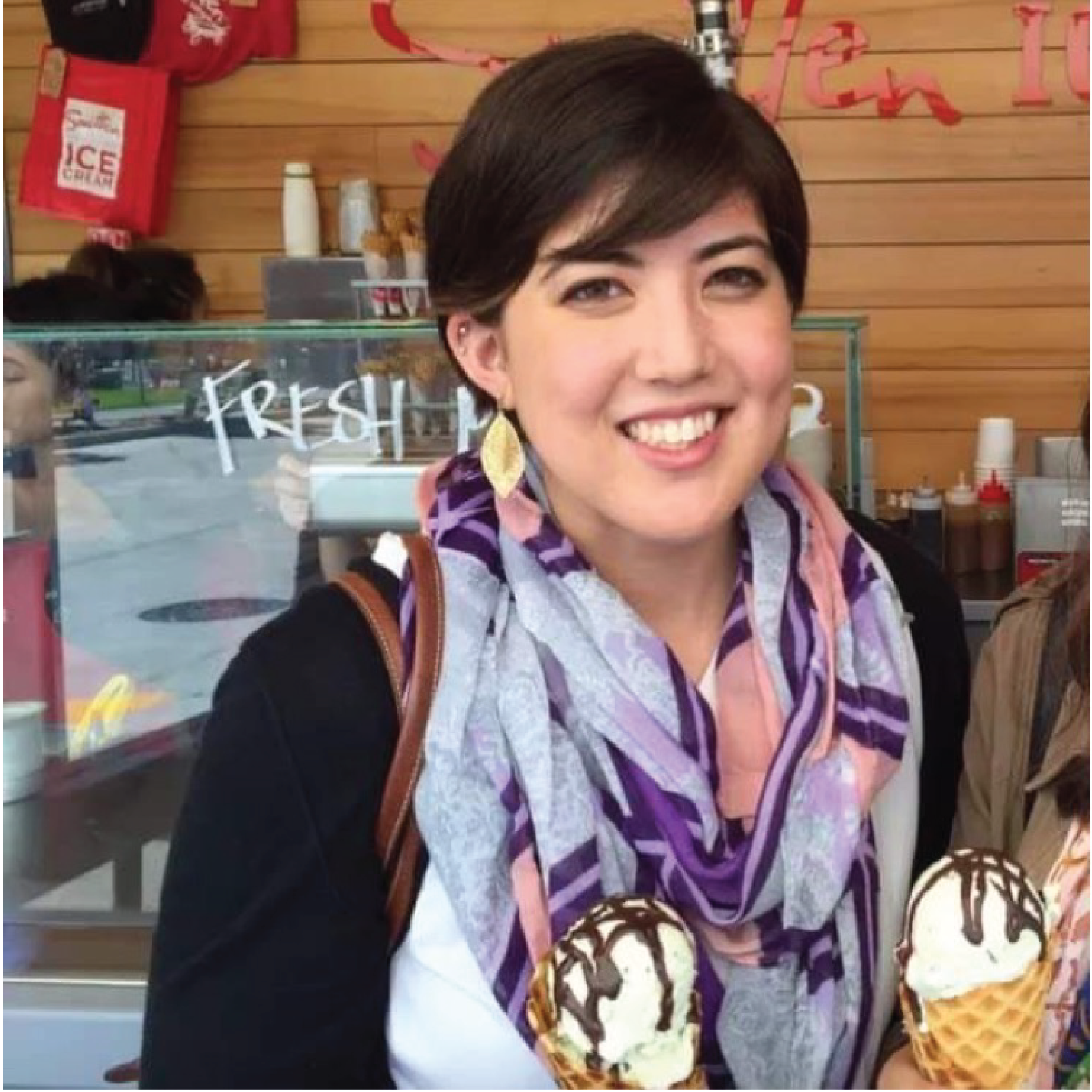 A feminine multiracial person in her mid-twenties is smiling and holding an ice cream cone. She has short brown hair and is wearing gold leaf-shaped earrings a scarf with a purple and pink striped and floral pattern.