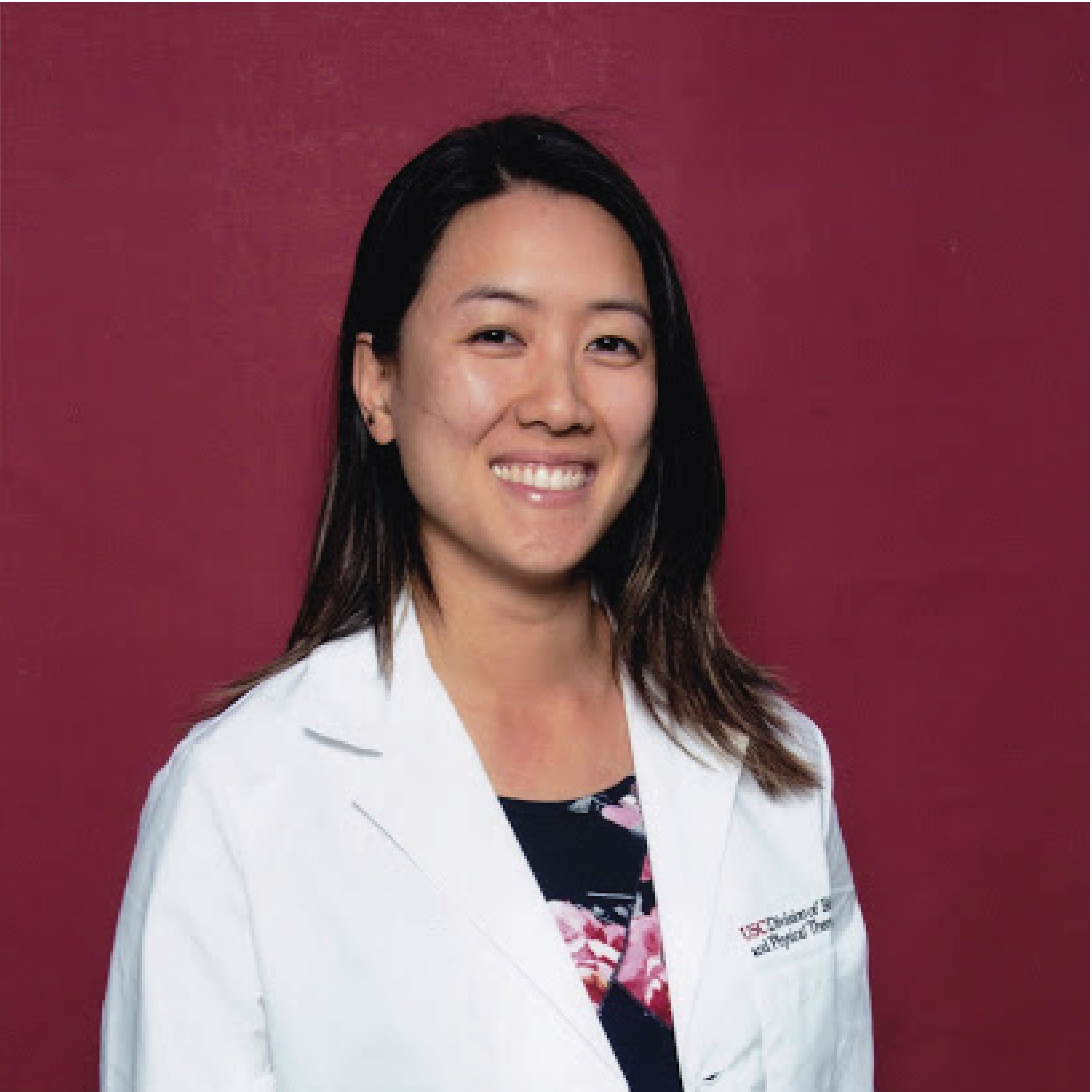 Portrait of Vivian, a long haired Asian woman wearing a white coat and floral dress in front of a solid red background. 
